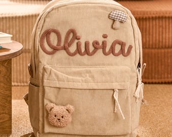 May include: Beige corduroy backpack with the name "Olivia" embroidered in brown thread. Features a small teddy bear patch and a mushroom applique. Includes side pockets and a front pocket with a zipper.