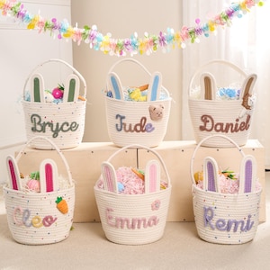 Peut inclure: Six paniers de Pâques en corde blanche avec des oreilles de lapin, chacun personnalisé avec un nom en fil coloré. Les paniers sont remplis d'herbe et d'œufs de Pâques colorés. Une guirlande décorative est suspendue au-dessus.