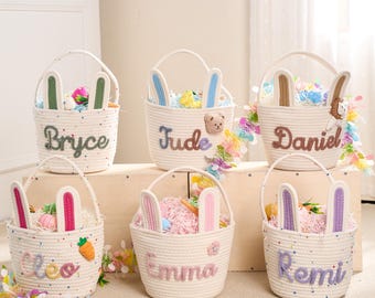 Può includere: Sei cestini pasquali bianchi in corda con orecchie da coniglio e nomi personalizzati. Ogni cestino è riempito con erba pasquale colorata e uova decorative. I nomi ricamati sui cestini sono Bryce, Jude, Daniel, Cleo, Emma e Remi.