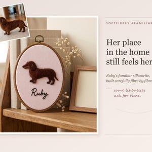 May include: An oval wooden embroidery hoop containing a light pink fabric background with a dark brown needle-felted dachshund silhouette and the name Ruby embroidered in black script. A small photo of a brown dachshund is attached to the top left.