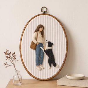 May include: An oval embroidery hoop wall hanging featuring a needle-felted scene of a person with long brown hair wearing a cream sweater and blue jeans, standing next to a black and white dog. The fabric background has vertical beige stripes.