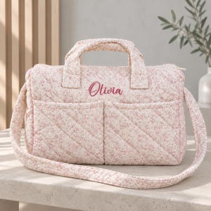 May include: A quilted fabric bag featuring a light pink and white floral pattern. The bag has two top handles, a long shoulder strap, and two front pockets. The name Olivia is embroidered in a dark pink script font on the front of the bag.