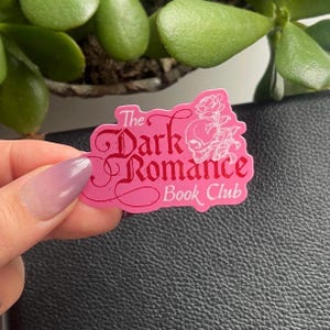 May include: Pink sticker with white text that reads "The Dark Romance Book Club". The sticker features a white skull with a rose design.