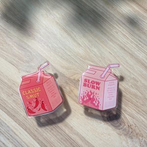 May include: Two clear plastic juice box stickers with pink and white text. The first sticker says "Classic Smut" with a red chili pepper design. The second sticker says "Slow Burn" with a red flame design.