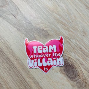 May include: A pink heart-shaped sticker with white text that reads "Team whoever the villain is".