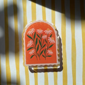 May include: A clear sticker with an orange background featuring white daisies with green stems and leaves. The sticker has a scalloped edge and is on a yellow and white striped background.