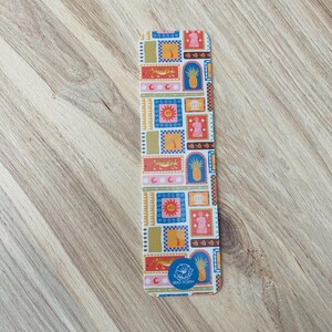 May include: A white bookmark with a colorful pattern of geometric shapes, pineapples, suns, and tigers. The bookmark has the text "Mad Poppy" in a blue circle at the bottom.