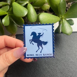 May include: Blue and white sticker with a silhouette of a cowboy riding a horse and lassoing. The text "Rebel Blue Ranch" is printed below the image.