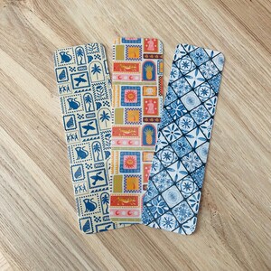 May include: Three colorful bookmarks with different patterns. The first bookmark has a white background with blue and yellow geometric shapes and animals. The second bookmark has a white background with orange, yellow, and blue geometric shapes and animals. The third bookmark has a white background with blue and white geometric shapes.