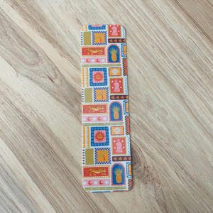 May include: A colorful bookmark with a repeating pattern of pineapples, tigers, suns, and other geometric shapes. The bookmark is white with a light blue, yellow, orange, and red color scheme.