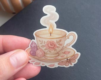 May include: A white teacup with a floral design and a lit candle inside, sitting on a saucer with pink and purple crystals and a brown moth.