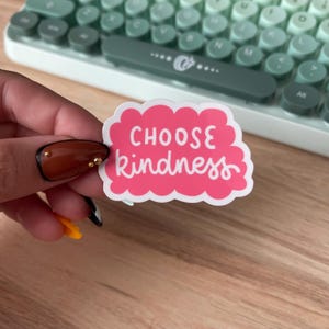 May include: A pink sticker with the words "CHOOSE kindness" in white script. The sticker is cloud-shaped and is held in front of a green and white keyboard.