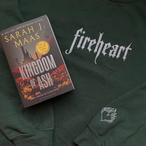 May include: A dark green sweatshirt with the words "fireheart" embroidered in silver on the front. The sweatshirt has a small silver heart with flames embroidered on the left sleeve. A book titled "Kingdom of Ash" by Sarah J. Maas is lying on top of the sweatshirt.