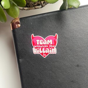 May include: Pink heart-shaped sticker with devil horns and the text "Team whoever the Villain is".
