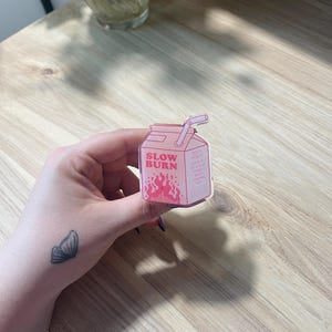 May include: Pink and white cartoon illustration of a milk carton with a straw. The carton has the words "Slow Burn" printed on it and a red flame design.
