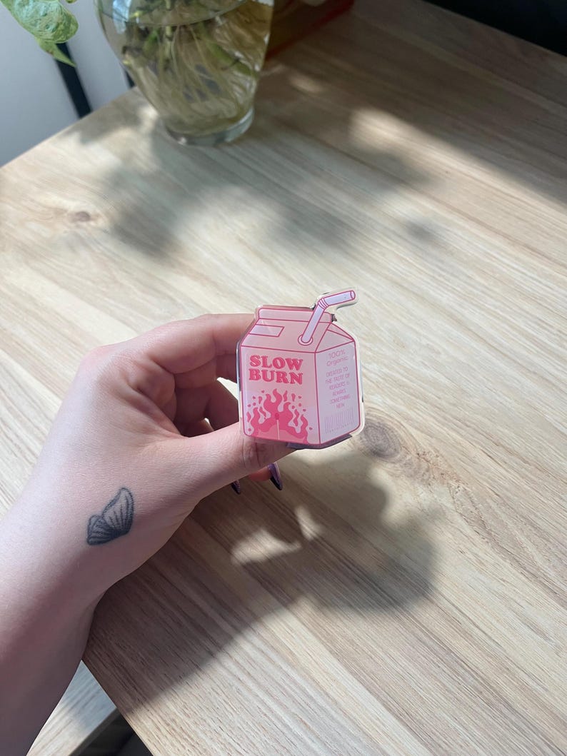 May include: Pink and white cartoon illustration of a milk carton with a straw. The carton has the words "Slow Burn" printed on it and a red flame design.