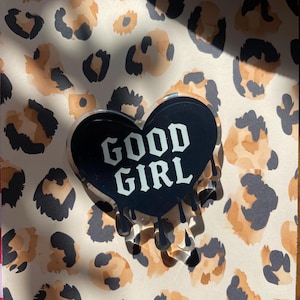 May include: A black heart-shaped pin with the words "GOOD GIRL" in white, dripping with black paint. The pin is set against a background of leopard print in shades of brown, black, and cream.