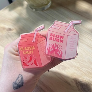 May include: Two pink and white cartoon juice boxes with straws. The juice boxes are clear and have a red and white design. One juice box says "Classic Smut" with red chili peppers. The other juice box says "Slow Burn" with red flames.