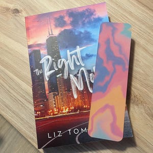 May include: A book titled "The Right Move" by Liz Tom with a colorful abstract bookmark. The book cover features a cityscape at sunset with a tall building in the center.