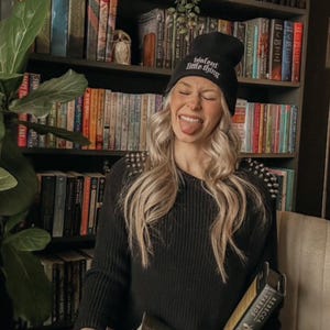 May include: A woman wearing a black beanie with the text "Valent little thing" is holding a stack of books. The books are all different colors and have different titles. The woman is sitting in a chair in front of a bookshelf filled with books.