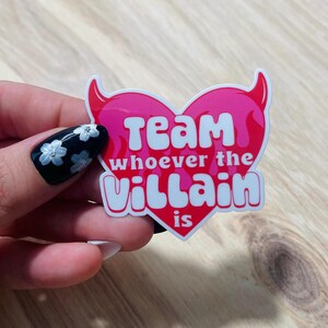 May include: A pink heart-shaped sticker with white text that reads "Team whoever the villain is". The heart has devil horns and flames on the sides.