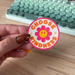 May include: A round sticker with the words "CHOOSE KINDNESS" in orange, surrounding a pink flower with a yellow smiley face center. The sticker is held in front of a mint green keyboard.