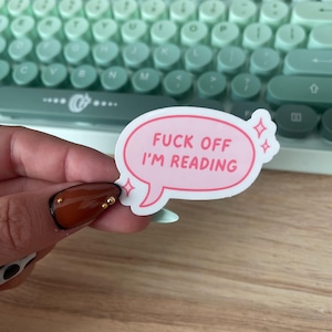 May include: A pink speech bubble sticker with the text "FUCK OFF I'M READING" in a bold, sans-serif font. The sticker has a white border and decorative starbursts. The background includes a mint green keyboard.