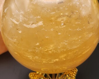May include: A large, clear, golden citrine crystal sphere on a gold butterfly stand. The sphere is smooth and polished, with a slight shimmer.