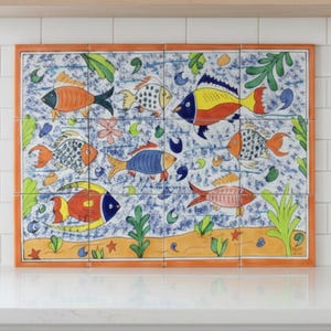 May include: A rectangular, hand-painted tile mural featuring colorful fish designs. The artwork showcases various fish in shades of orange, yellow, blue, and red, set against a blue and white background. The mural is framed with an orange border and includes floral and aquatic elements.