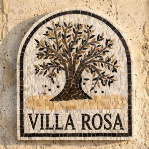 May include: An arched stone mosaic wall plaque featuring a central olive tree design with dark brown and gold leaves. The text VILLA ROSA is displayed in bold black capital letters at the bottom, framed by a black border on a white background.