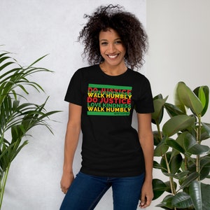 May include: Black t-shirt with the text "DO JUSTICE WALK HUMBLY DO JUSTICE LOVE KINDNESS WALK HUMBLY WITH YOUR GOD" in green, red, and yellow. The shirt is worn by a person in front of a green leafy background.