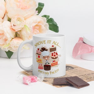 Könnte beinhalten: Eine weiße Keramiktasse mit einem goldenen Design, das einen Cartoon-Bäcker und verschiedene Backwaren zeigt. Der Text auf der Tasse lautet "Shove it all... into my face!!"
