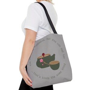 May include: A gray tote bag with a black strap and a design of a green teapot and a teacup with a pink flower. The text on the bag reads "You can't buy happiness, but you can buy tea...that's kinda the same thing."