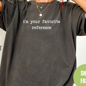 Könnte beinhalten: Ein dunkelgraues Oversize-T-Shirt mit dem Aufdruck "i'm your favorite reference" in Weiß.