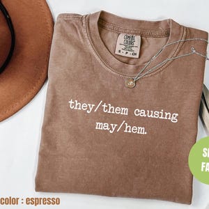 Könnte beinhalten: Espresso-braunes T-Shirt mit dem in Weiß gedruckten Text "they/them causing may/hem."