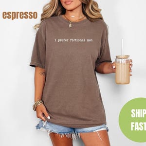 Pu&ograve; includere: T-shirt color espresso con la scritta "I prefer fictional men" in un font semplice. La maglietta &egrave; abbinata a pantaloncini di jeans. Una persona tiene in mano un bicchiere con una cannuccia e un cerchio verde con la scritta "SHIPS FAST!"