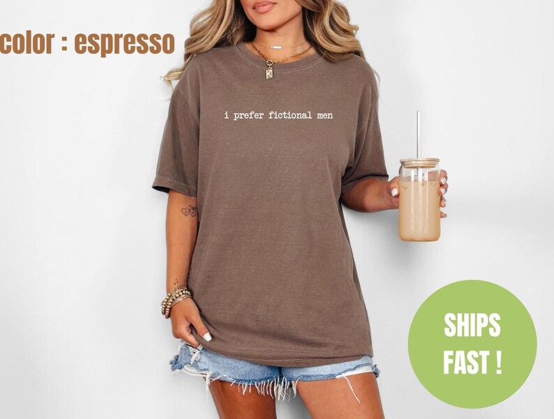 Pu&ograve; includere: T-shirt color espresso con la scritta "I prefer fictional men" in un font semplice. La maglietta &egrave; abbinata a pantaloncini di jeans. Una persona tiene in mano un bicchiere con una cannuccia e un cerchio verde con la scritta "SHIPS FAST!"