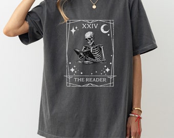 Pode incluir: Uma camiseta cinza escura de tamanho grande com um gráfico branco de uma carta de tarô. O cartão mostra um esqueleto lendo um livro com o texto "XXIV THE READER" e uma lua crescente.