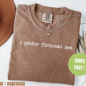 Pu&ograve; includere: T-shirt marrone con la scritta bianca "i prefer fictional men". La maglietta ha un girocollo e un'etichetta Comfort Colors. Una collana d'argento con un ciondolo &egrave; drappeggiata sulla maglietta.