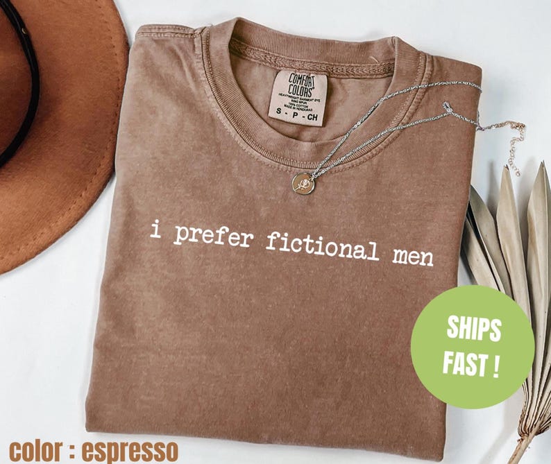 Pu&ograve; includere: T-shirt marrone con la scritta bianca "i prefer fictional men". La maglietta ha un girocollo e un'etichetta Comfort Colors. Una collana d'argento con un ciondolo &egrave; drappeggiata sulla maglietta.