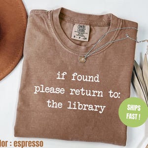 May include: Espresso-colored t-shirt with the text "if found please return to: the library" in white. The shirt has a crew neck and a small tag at the neckline. A silver necklace with a pendant is draped across the shirt.