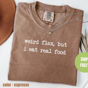 Puede incluir: Camiseta marrón espresso con el texto "weird flex, but i eat real food" impreso en blanco.