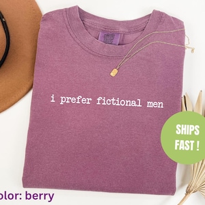Pu&ograve; includere: T-shirt color bacca con la scritta "i prefer fictional men" stampata in bianco. Una collana d'oro &egrave; appoggiata sulla maglietta. La maglietta &egrave; piegata e ha un'etichetta Comfort Colors. Un cerchio verde con "SHIPS FAST!" &egrave; visibile.