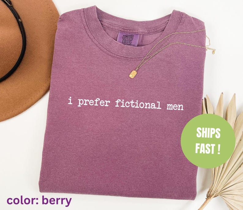 Pu&ograve; includere: T-shirt color bacca con la scritta "i prefer fictional men" stampata in bianco. Una collana d'oro &egrave; appoggiata sulla maglietta. La maglietta &egrave; piegata e ha un'etichetta Comfort Colors. Un cerchio verde con "SHIPS FAST!" &egrave; visibile.