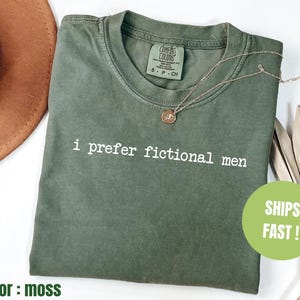 Pu&ograve; includere: T-shirt verde muschio con la scritta bianca "i prefer fictional men". La maglietta ha un girocollo e maniche corte. Una collana d'oro con un ciondolo &egrave; sulla maglietta. La maglietta &egrave; piegata su una superficie bianca.