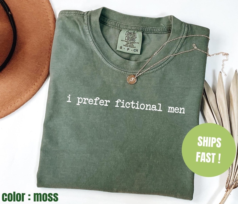 Pu&ograve; includere: T-shirt verde muschio con la scritta bianca "i prefer fictional men". La maglietta ha un girocollo e maniche corte. Una collana d'oro con un ciondolo &egrave; sulla maglietta. La maglietta &egrave; piegata su una superficie bianca.