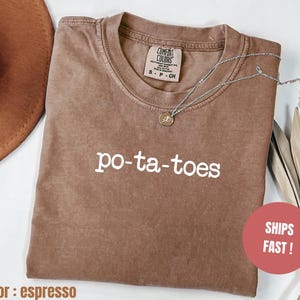 Op de afbeelding: Espresso-kleurig T-shirt met het woord "po-ta-toes" in het wit gedrukt. Het shirt heeft een ronde hals en korte mouwen. Een zilveren ketting met een hanger is over het shirt gedrapeerd. Het shirt is gemaakt van een zacht materiaal.