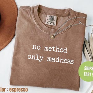 May include: Espresso-colored t-shirt with the phrase "no method only madness" in white. The shirt has a crew neck and a small tag at the neckline. A silver necklace with a pendant is draped across the shirt. The shirt is a size small.