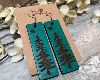 Könnte beinhalten: Rechteckige, türkisfarbene Holzohrringe mit einem dunkelbraunen, lasergravierten Tannenbaum und einem kleinen Stern an der Spitze. Die Ohrringe sind an einer hellbraunen Pappkarte mit dem aufgedruckten Text 4 The Love of Wood befestigt.