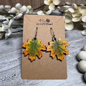 May include: A pair of wooden leaf earrings with green, yellow, and orange coloring. The earrings are on a brown card with the text "4 The Love Of Wood". The earrings are hanging from bronze-colored hooks.